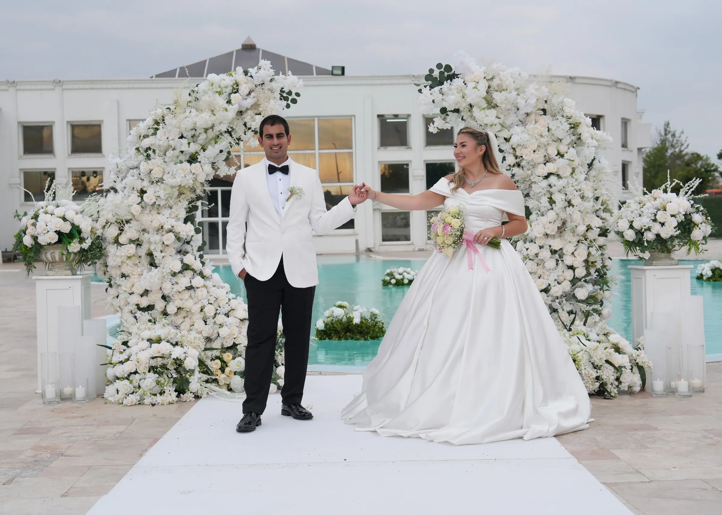Beyaz çiçekli kemer altında el ele tutuşan gelin ve damat, arkada havuz ve beyaz köşk